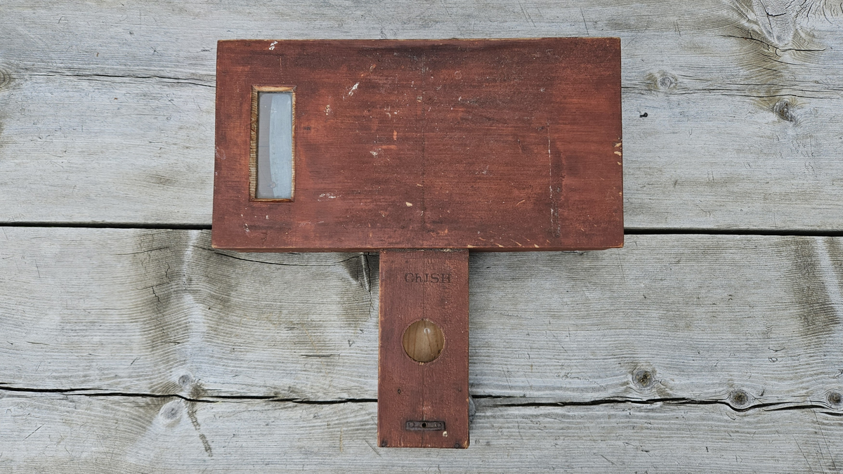 Nivelleringsinstrument bestående av trekasse med glassvindu for avlesing av mål. Fra skiva med mål henger et lodd. På baksida av trekassa er en bøyle; sannsynligvis for feste på et stativ.