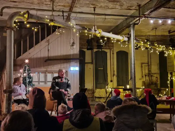 Personer på scenen og publikum med nisseluer i industrilokale med julelys i taket.