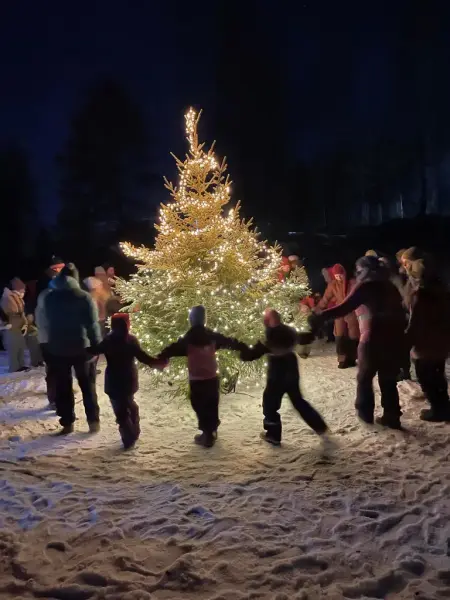 Barn holder hender og går rundt et grantre med lys i mørke omgivelser