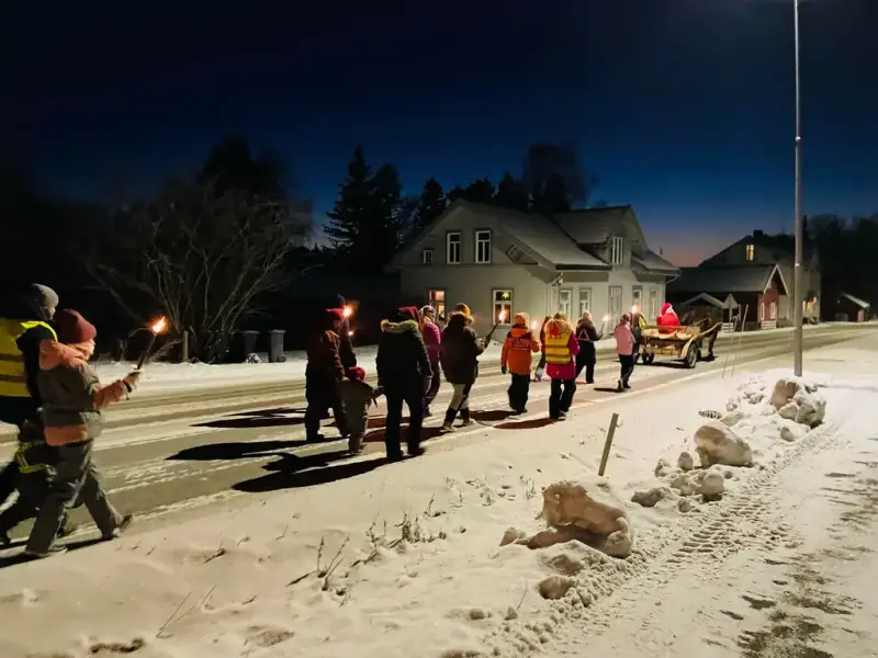 Mørkt og snø med gatelys, personer går i fakkeltog i boligstrøk. Nisse med hest og kjerre går først.