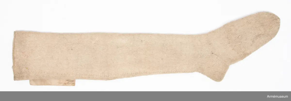 Lång vit ullstrumpa.

Strumpan är slätstickad av vitt ullgarn och lätt valkad.
Skaftet är avslutat med en enkel rullkant. Hälen är rak. Från foten och upp löper en "söm" på baksiden av vaden. Denna ser ut att vara stickad med varannat varv räta och aviga maskor.
Strumpan är märkt med röda vaxsigill på skaftet och vid tån.
På den samhörande modelletiketten av gulnat papper finns följande text:
"Af Kongl. Majt Nådigast faststäldt Modell å Ullstrumpor för Manskapet af alla Vapen, enl. General Order af d. 20. Maji 1826.
På Befallning, T. W. Lode, Adjut. i Kgl. Krigs Colleg."