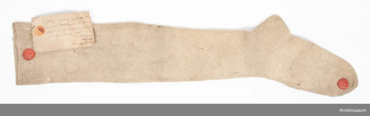 Lång vit ullstrumpa.

Strumpan är slätstickad av vitt ullgarn och lätt valkad.
Skaftet är avslutat med en enkel rullkant. Hälen är rak. Från foten och upp löper en "söm" på baksiden av vaden. Denna ser ut att vara stickad med varannat varv räta och aviga maskor.
Strumpan är märkt med röda vaxsigill på skaftet och vid tån.
På den samhörande modelletiketten av gulnat papper finns följande text:
"Af Kongl. Majt Nådigast faststäldt Modell å Ullstrumpor för Manskapet af alla Vapen, enl. General Order af d. 20. Maji 1826.
På Befallning, T. W. Lode, Adjut. i Kgl. Krigs Colleg."