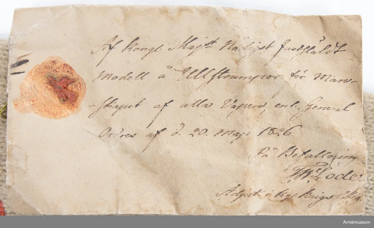 Lång vit ullstrumpa.

Strumpan är slätstickad av vitt ullgarn och lätt valkad.
Skaftet är avslutat med en enkel rullkant. Hälen är rak. Från foten och upp löper en "söm" på baksiden av vaden. Denna ser ut att vara stickad med varannat varv räta och aviga maskor.
Strumpan är märkt med röda vaxsigill på skaftet och vid tån.
På den samhörande modelletiketten av gulnat papper finns följande text:
"Af Kongl. Majt Nådigast faststäldt Modell å Ullstrumpor för Manskapet af alla Vapen, enl. General Order af d. 20. Maji 1826.
På Befallning, T. W. Lode, Adjut. i Kgl. Krigs Colleg."