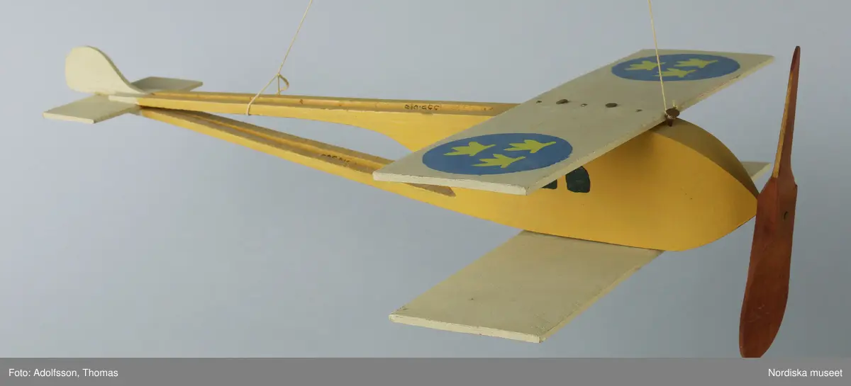 Flygplansmodell av trä, med dubbla vingar och propeller. Delarna är ihopspikade. Målad i lejongult, på vingarna beiga med blå cirklar med tre kronor i ljusgult och röd propeller. Bomullssnöre för upphängning. Har använts som prydnad i taket. Flygplanets propeller är skadat i kanten och de rostiga spikarna behöver tryckas in.