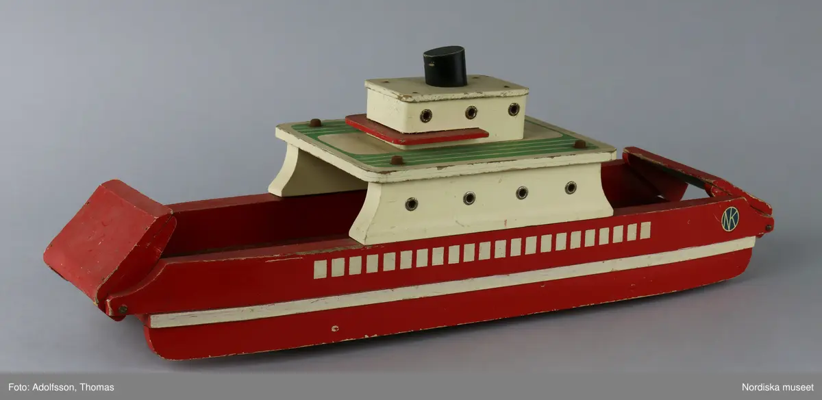 Båt av trä, bilfärja med uppfällbara ramper. Målad i rött, vitt, grönt och svart. Färgen delvis flagnad. Delarna hålls ihop av skruvar och spikar som rostat. Golvet är beklätt med en grön "matta" av papper. Färjan är märkt med NK:s logga på två etiketter som sitter fast på båda sidorna av fören. På undersidan finns fyra gummihjul. Mellan hjulen har någon klottrat namn med barnslig stil: troligen "ERIK" och "NK". I mitten står ett namn som är svårt att tyda.