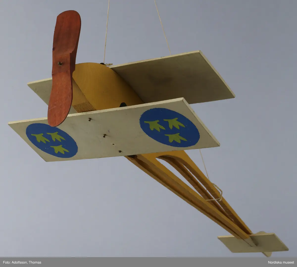 Flygplansmodell av trä, med dubbla vingar och propeller. Delarna är ihopspikade. Målad i lejongult, på vingarna beiga med blå cirklar med tre kronor i ljusgult och röd propeller. Bomullssnöre för upphängning. Har använts som prydnad i taket. Flygplanets propeller är skadat i kanten och de rostiga spikarna behöver tryckas in.