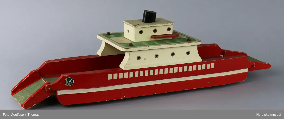 Båt av trä, bilfärja med uppfällbara ramper. Målad i rött, vitt, grönt och svart. Färgen delvis flagnad. Delarna hålls ihop av skruvar och spikar som rostat. Golvet är beklätt med en grön "matta" av papper. Färjan är märkt med NK:s logga på två etiketter som sitter fast på båda sidorna av fören. På undersidan finns fyra gummihjul. Mellan hjulen har någon klottrat namn med barnslig stil: troligen "ERIK" och "NK". I mitten står ett namn som är svårt att tyda.