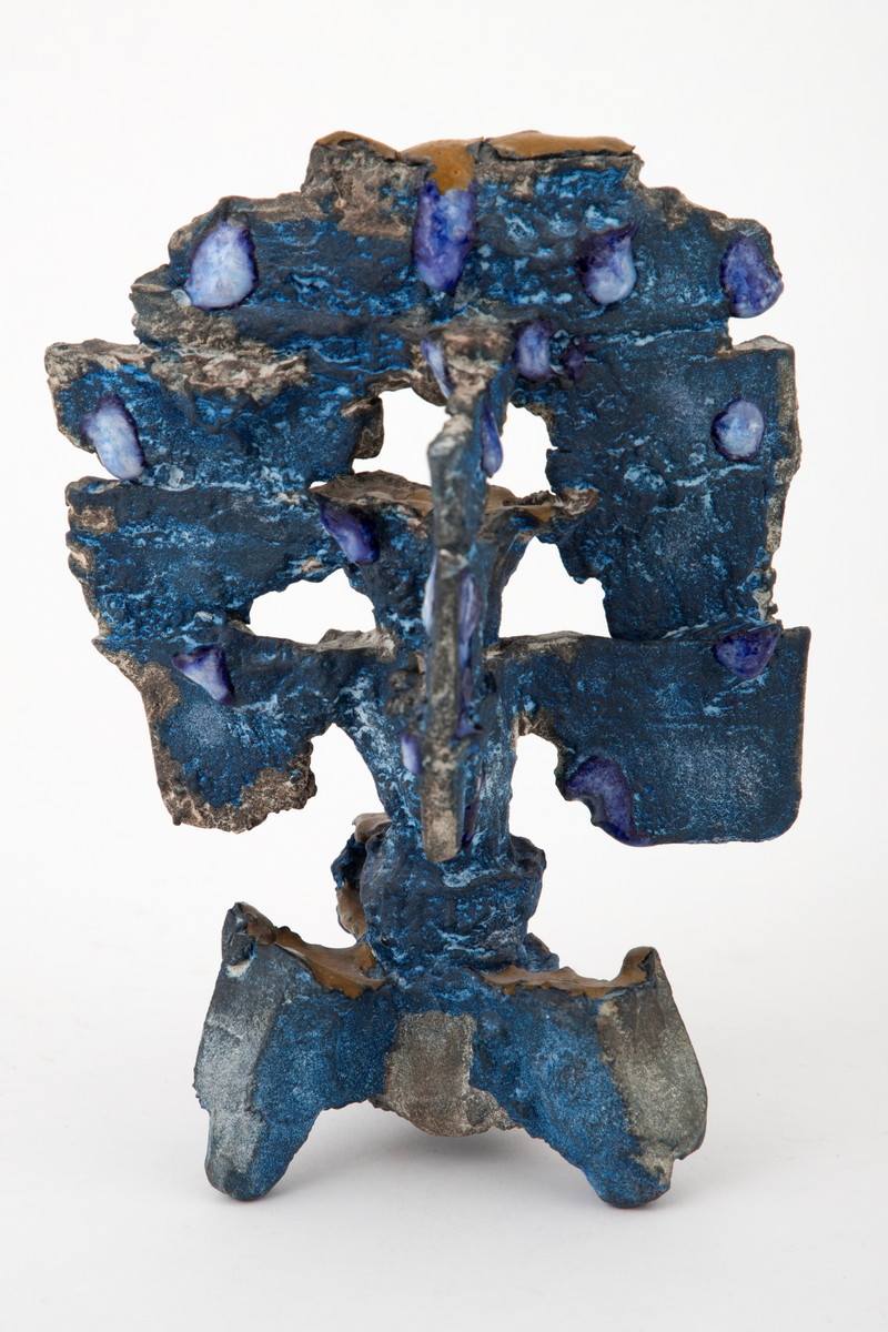 Abstrakt keramisk objekt med en oppstående form på tre grovt utformede føtter. Toppstykket har en vertikal utforming og er delvis gjennombrutt. Overflaten er ru og farget blå. Delvis dekket med dråpeformede glasurpletter i blått og hvitt fordelt på skulpturen. Noen små horisontale flater er dekket med okergul glasur.