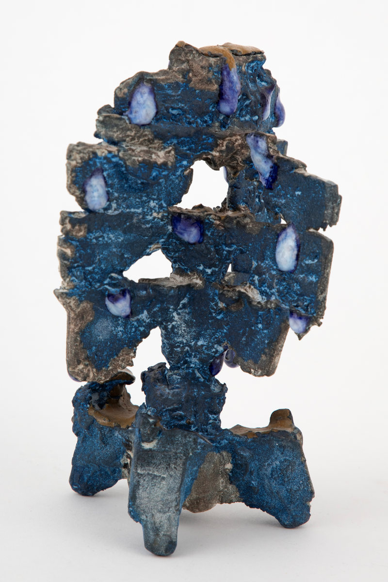 Abstrakt keramisk objekt med en oppstående form på tre grovt utformede føtter. Toppstykket har en vertikal utforming og er delvis gjennombrutt. Overflaten er ru og farget blå. Delvis dekket med dråpeformede glasurpletter i blått og hvitt fordelt på skulpturen. Noen små horisontale flater er dekket med okergul glasur.