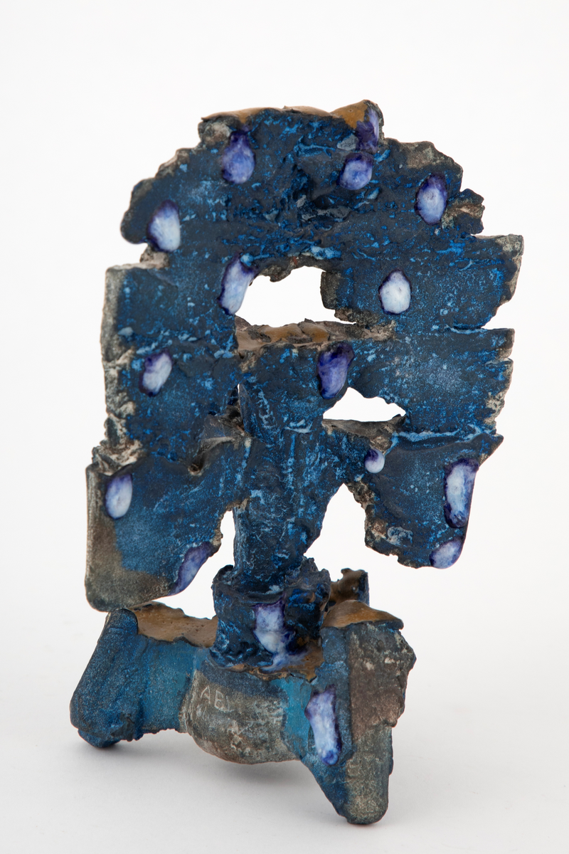 Abstrakt keramisk objekt med en oppstående form på tre grovt utformede føtter. Toppstykket har en vertikal utforming og er delvis gjennombrutt. Overflaten er ru og farget blå. Delvis dekket med dråpeformede glasurpletter i blått og hvitt fordelt på skulpturen. Noen små horisontale flater er dekket med okergul glasur.