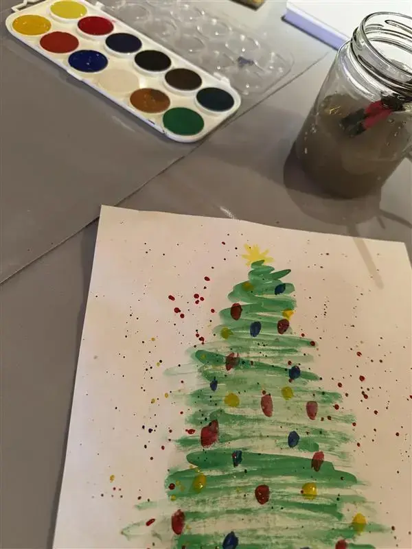 Vi ser vannfarger, et glass med skitten vann og malerkoster og en fin akvarell av et juletre med fargede lys