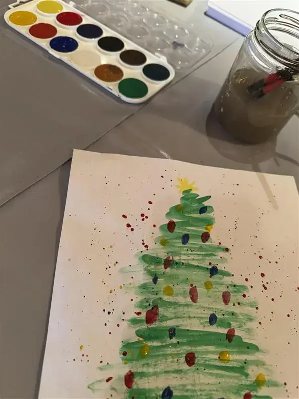 Vi ser vannfarger, et glass med skitten vann og malerkoster og en fin akvarell av et juletre med fargede lys