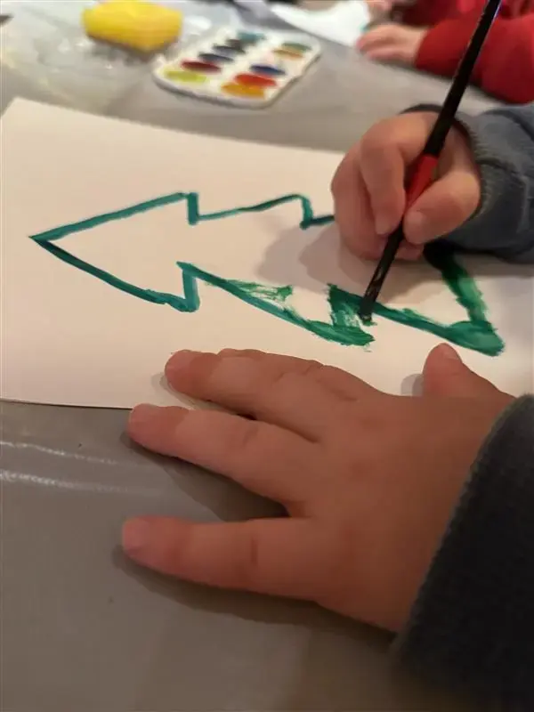 Vi ser en barnehånd som maler et grønt juletre på et hvitt ark