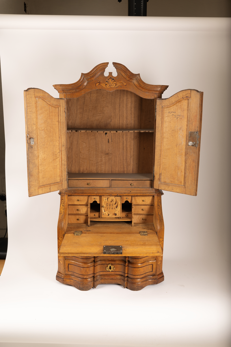 Barneskatoll i rokokkostil med separat overskap og underskap samt felles nøkkel til begge deler. Underskap med kraftig buet og profilert front, to skuffer samt nedfellbart pultlokk med hylle-, skap- og skuffseksjon innenfor. Overskap med to rette dører og kraftig profilert, delvis gjennombrutt gesims. Både over- og underskapet rikt dekorert med intarsia av høy kvalitet med rocailler, skjell, konturlinjer og blomsterornamentikk. Dekorative skilt av messing rundt alle nøkkelhull. Initialer og årstall på skapdør bak pultlokk. Skatollet står på to lave føtter.

A: underskap
B: nøkkel
C: overskap