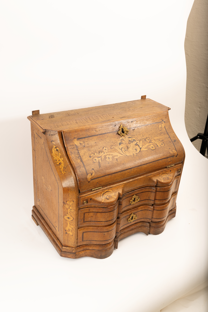 Barneskatoll i rokokkostil med separat overskap og underskap samt felles nøkkel til begge deler. Underskap med kraftig buet og profilert front, to skuffer samt nedfellbart pultlokk med hylle-, skap- og skuffseksjon innenfor. Overskap med to rette dører og kraftig profilert, delvis gjennombrutt gesims. Både over- og underskapet rikt dekorert med intarsia av høy kvalitet med rocailler, skjell, konturlinjer og blomsterornamentikk. Dekorative skilt av messing rundt alle nøkkelhull. Initialer og årstall på skapdør bak pultlokk. Skatollet står på to lave føtter.

A: underskap
B: nøkkel
C: overskap