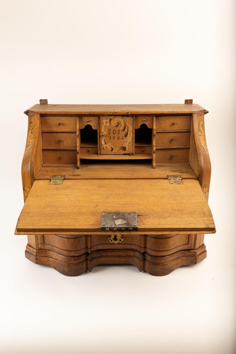 Barneskatoll i rokokkostil med separat overskap og underskap samt felles nøkkel til begge deler. Underskap med kraftig buet og profilert front, to skuffer samt nedfellbart pultlokk med hylle-, skap- og skuffseksjon innenfor. Overskap med to rette dører og kraftig profilert, delvis gjennombrutt gesims. Både over- og underskapet rikt dekorert med intarsia av høy kvalitet med rocailler, skjell, konturlinjer og blomsterornamentikk. Dekorative skilt av messing rundt alle nøkkelhull. Initialer og årstall på skapdør bak pultlokk. Skatollet står på to lave føtter.

A: underskap
B: nøkkel
C: overskap