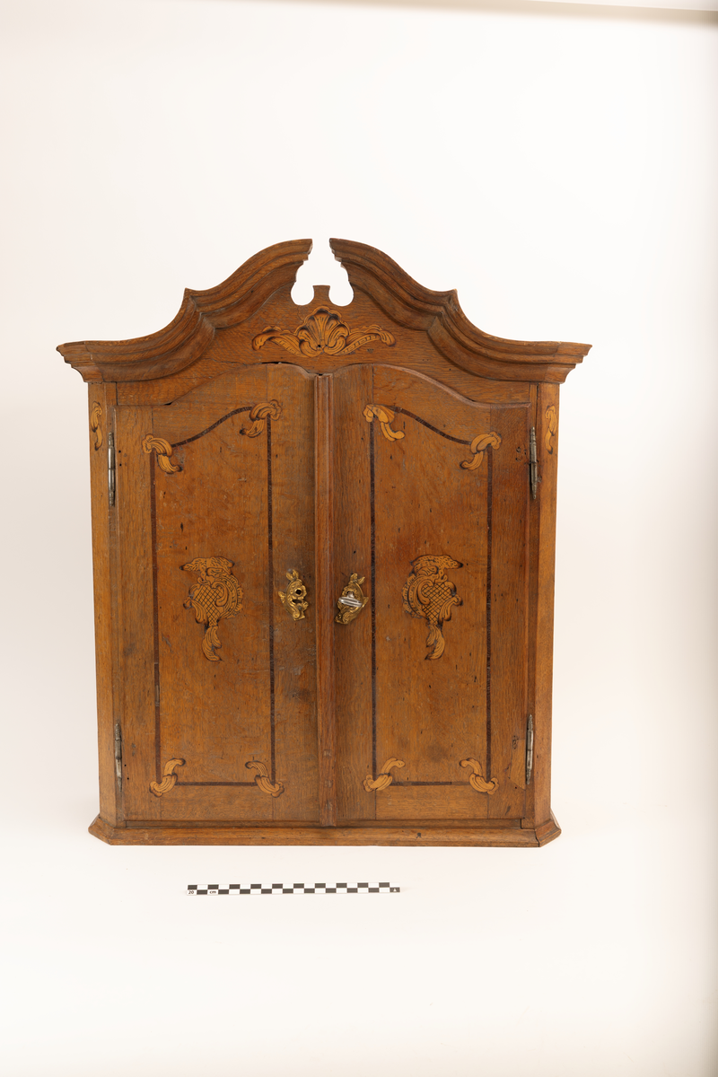 Barneskatoll i rokokkostil med separat overskap og underskap samt felles nøkkel til begge deler. Underskap med kraftig buet og profilert front, to skuffer samt nedfellbart pultlokk med hylle-, skap- og skuffseksjon innenfor. Overskap med to rette dører og kraftig profilert, delvis gjennombrutt gesims. Både over- og underskapet rikt dekorert med intarsia av høy kvalitet med rocailler, skjell, konturlinjer og blomsterornamentikk. Dekorative skilt av messing rundt alle nøkkelhull. Initialer og årstall på skapdør bak pultlokk. Skatollet står på to lave føtter.

A: underskap
B: nøkkel
C: overskap