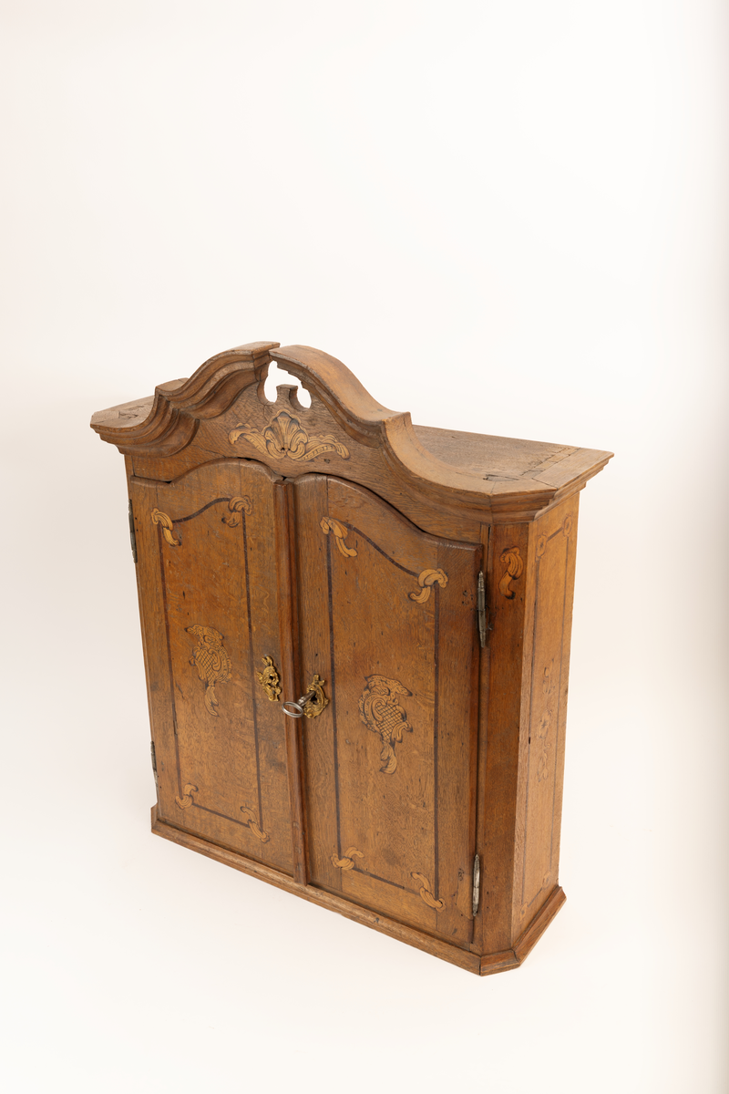 Barneskatoll i rokokkostil med separat overskap og underskap samt felles nøkkel til begge deler. Underskap med kraftig buet og profilert front, to skuffer samt nedfellbart pultlokk med hylle-, skap- og skuffseksjon innenfor. Overskap med to rette dører og kraftig profilert, delvis gjennombrutt gesims. Både over- og underskapet rikt dekorert med intarsia av høy kvalitet med rocailler, skjell, konturlinjer og blomsterornamentikk. Dekorative skilt av messing rundt alle nøkkelhull. Initialer og årstall på skapdør bak pultlokk. Skatollet står på to lave føtter.

A: underskap
B: nøkkel
C: overskap