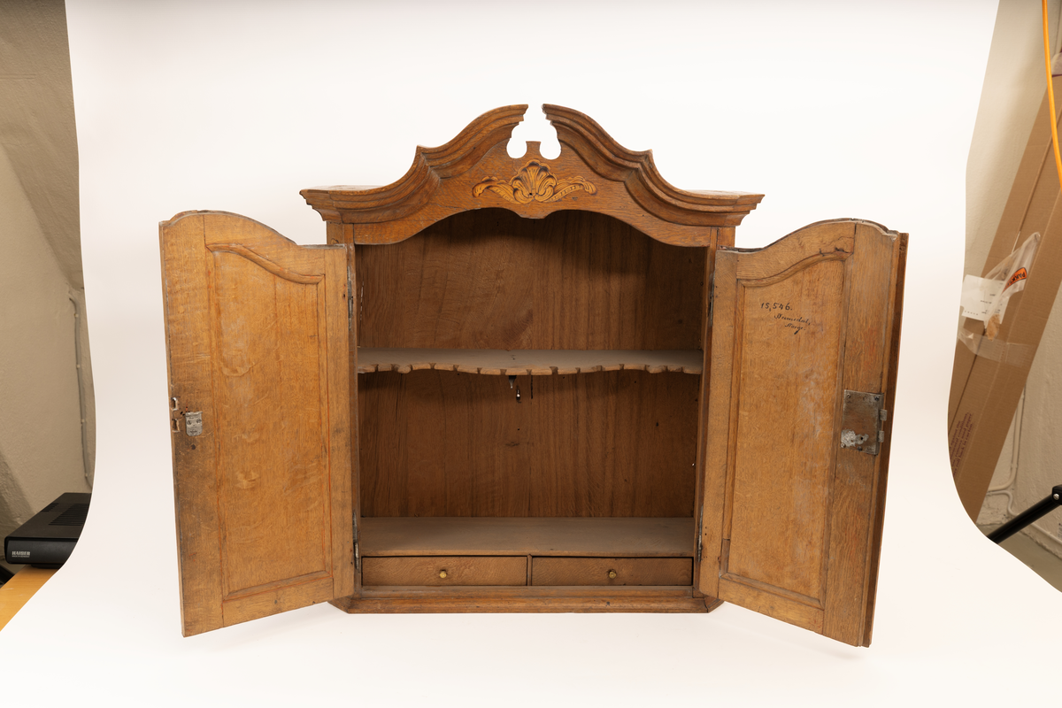 Barneskatoll i rokokkostil med separat overskap og underskap samt felles nøkkel til begge deler. Underskap med kraftig buet og profilert front, to skuffer samt nedfellbart pultlokk med hylle-, skap- og skuffseksjon innenfor. Overskap med to rette dører og kraftig profilert, delvis gjennombrutt gesims. Både over- og underskapet rikt dekorert med intarsia av høy kvalitet med rocailler, skjell, konturlinjer og blomsterornamentikk. Dekorative skilt av messing rundt alle nøkkelhull. Initialer og årstall på skapdør bak pultlokk. Skatollet står på to lave føtter.

A: underskap
B: nøkkel
C: overskap