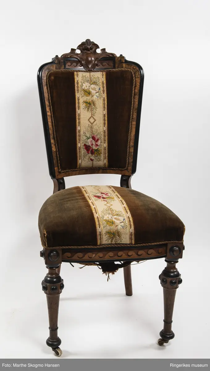 Stol i brun mahogni med utskjæringer. Hjul på frembeina. Brunt fløyelstrekk på setet med korsstingsbroderi av blomster og geometriske mønster.