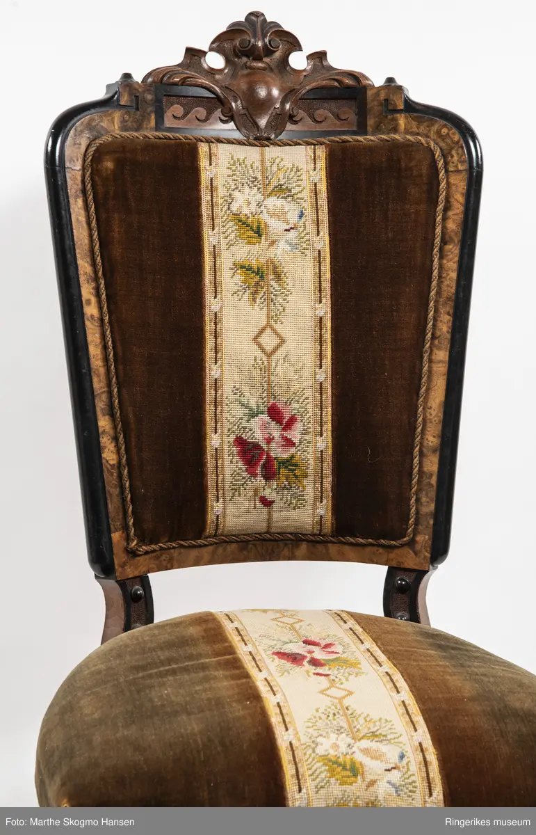 Stol i brun mahogni med utskjæringer. Hjul på frembeina. Brunt fløyelstrekk på setet med korsstingsbroderi av blomster og geometriske mønster.