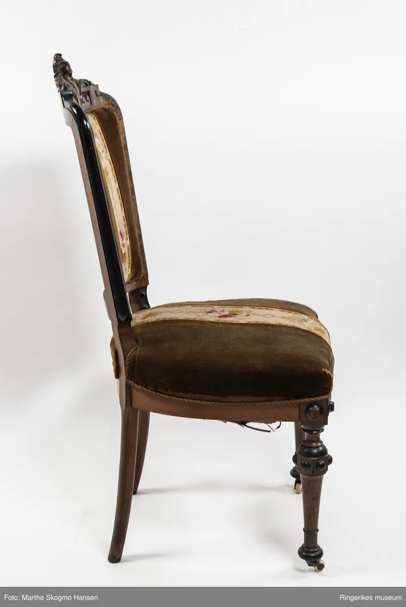 Stol i brun mahogni med utskjæringer. Hjul på frembeina. Brunt fløyelstrekk på setet med korsstingsbroderi av blomster og geometriske mønster.