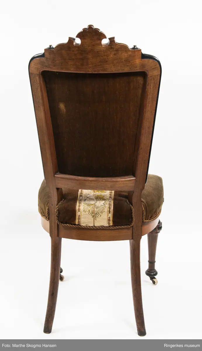 Stol i brun mahogni med utskjæringer. Hjul på frembeina. Brunt fløyelstrekk på setet med korsstingsbroderi av blomster og geometriske mønster.