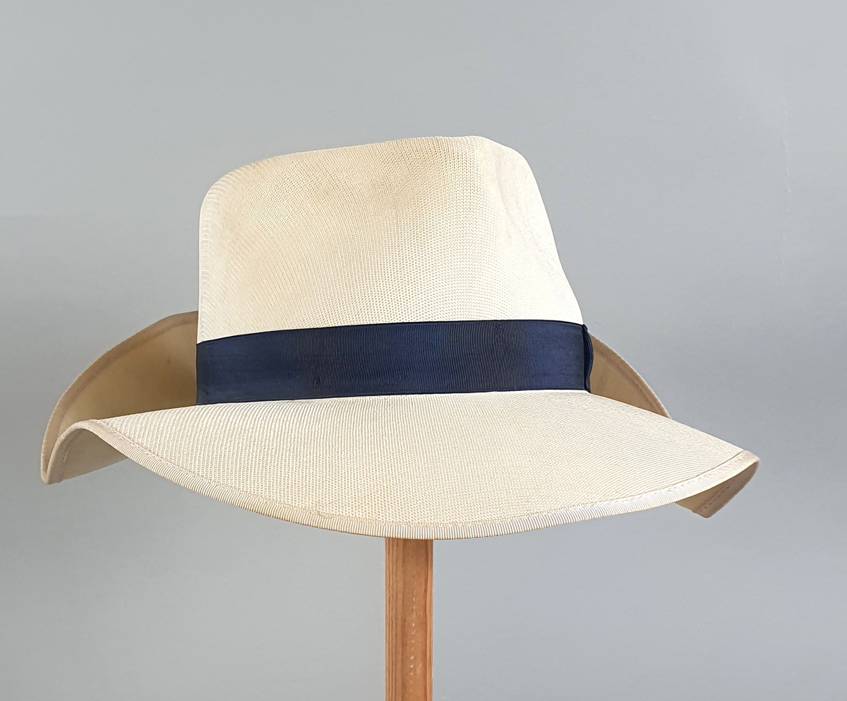 Hvit herrehatt med mørkeblått bånd rundt pullen. Bremmen er brettet opp bak. Svettebånd av skinn på innsiden av pullen med en liten hvit sløyfe midt bak.