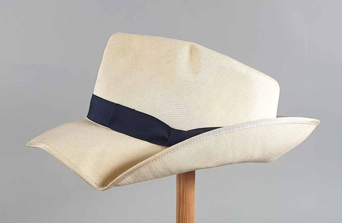 Hvit herrehatt med mørkeblått bånd rundt pullen. Bremmen er brettet opp bak. Svettebånd av skinn på innsiden av pullen med en liten hvit sløyfe midt bak.