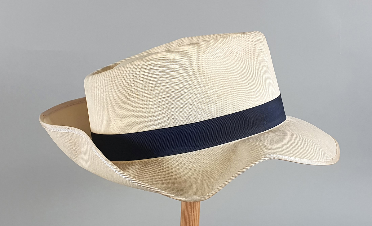 Hvit herrehatt med mørkeblått bånd rundt pullen. Bremmen er brettet opp bak. Svettebånd av skinn på innsiden av pullen med en liten hvit sløyfe midt bak.