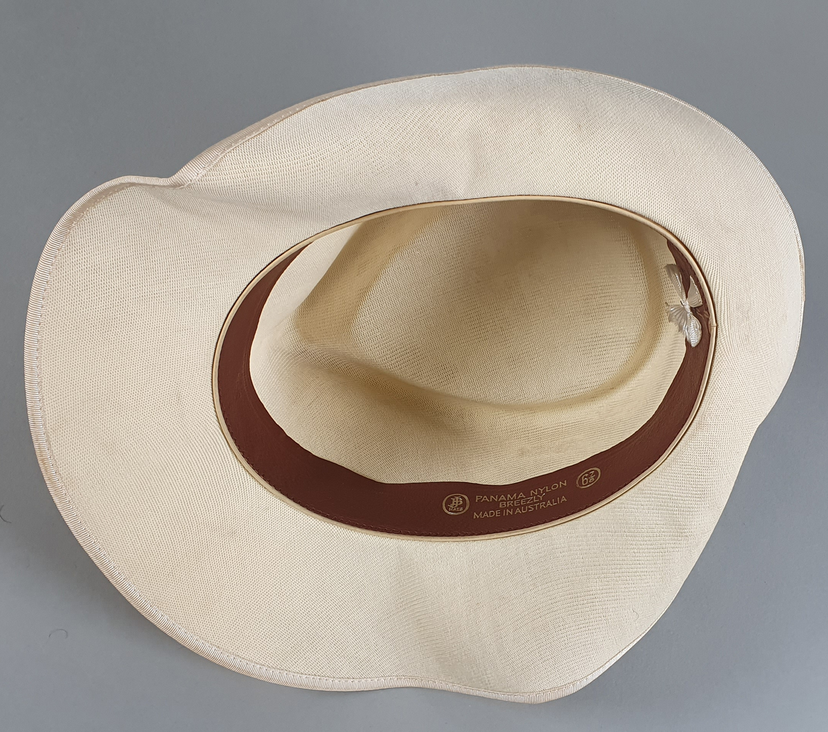 Hvit herrehatt med mørkeblått bånd rundt pullen. Bremmen er brettet opp bak. Svettebånd av skinn på innsiden av pullen med en liten hvit sløyfe midt bak.