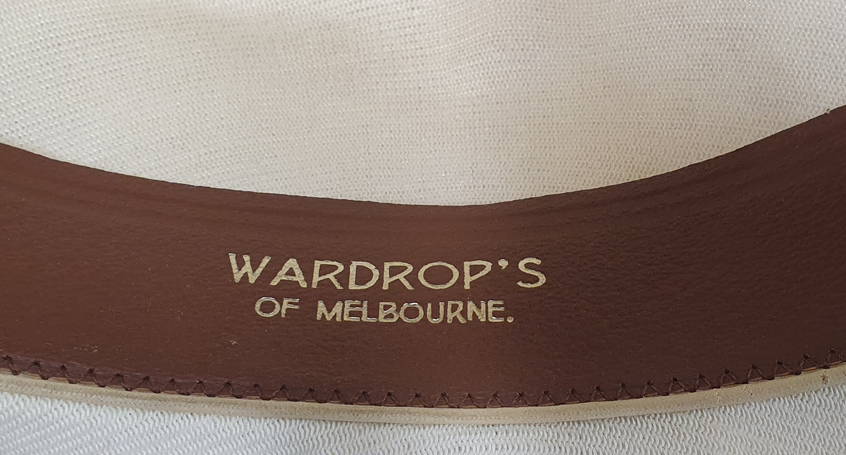 Hvit herrehatt med mørkeblått bånd rundt pullen. Bremmen er brettet opp bak. Svettebånd av skinn på innsiden av pullen med en liten hvit sløyfe midt bak.