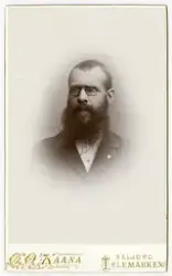 Portrett av mann med skjegg og briller. På jakken er det fes