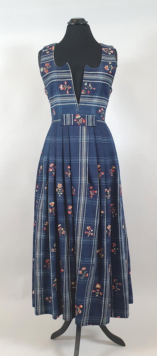 Blå stakk med liv. Hvite og lyseblå smale ruter på mørkeblå bunn, med broderte blomster spredt utover hele stakken og livet. Alle blomstene er ulike, brodert på frihånd. Livet er sydd på stakken i senere tid, og er fôret med hvitt bomullsstoff. Blå skoning av bomull på innsiden av skjørtekanten.