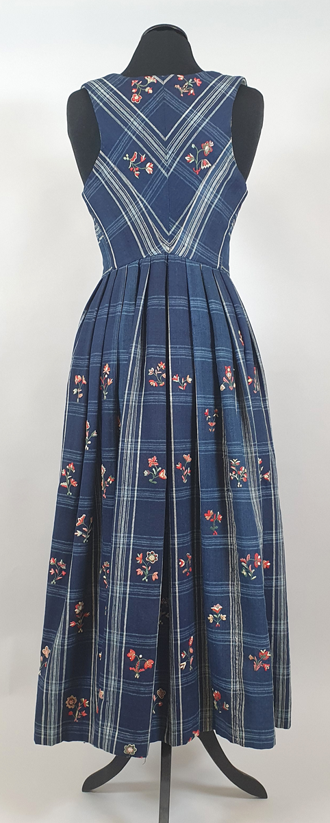 Blå stakk med liv. Hvite og lyseblå smale ruter på mørkeblå bunn, med broderte blomster spredt utover hele stakken og livet. Alle blomstene er ulike, brodert på frihånd. Livet er sydd på stakken i senere tid, og er fôret med hvitt bomullsstoff. Blå skoning av bomull på innsiden av skjørtekanten.