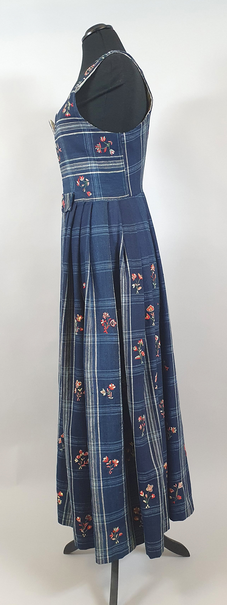 Blå stakk med liv. Hvite og lyseblå smale ruter på mørkeblå bunn, med broderte blomster spredt utover hele stakken og livet. Alle blomstene er ulike, brodert på frihånd. Livet er sydd på stakken i senere tid, og er fôret med hvitt bomullsstoff. Blå skoning av bomull på innsiden av skjørtekanten.