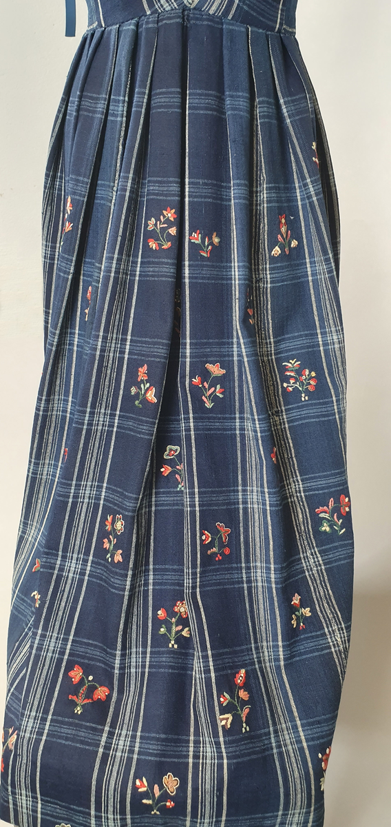 Blå stakk med liv. Hvite og lyseblå smale ruter på mørkeblå bunn, med broderte blomster spredt utover hele stakken og livet. Alle blomstene er ulike, brodert på frihånd. Livet er sydd på stakken i senere tid, og er fôret med hvitt bomullsstoff. Blå skoning av bomull på innsiden av skjørtekanten.