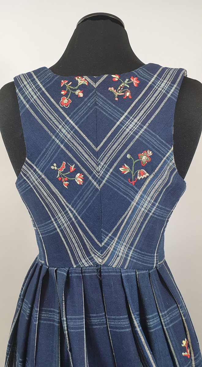 Blå stakk med liv. Hvite og lyseblå smale ruter på mørkeblå bunn, med broderte blomster spredt utover hele stakken og livet. Alle blomstene er ulike, brodert på frihånd. Livet er sydd på stakken i senere tid, og er fôret med hvitt bomullsstoff. Blå skoning av bomull på innsiden av skjørtekanten.