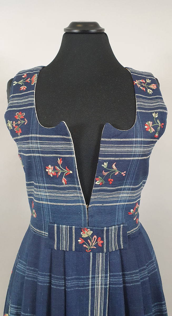 Blå stakk med liv. Hvite og lyseblå smale ruter på mørkeblå bunn, med broderte blomster spredt utover hele stakken og livet. Alle blomstene er ulike, brodert på frihånd. Livet er sydd på stakken i senere tid, og er fôret med hvitt bomullsstoff. Blå skoning av bomull på innsiden av skjørtekanten.