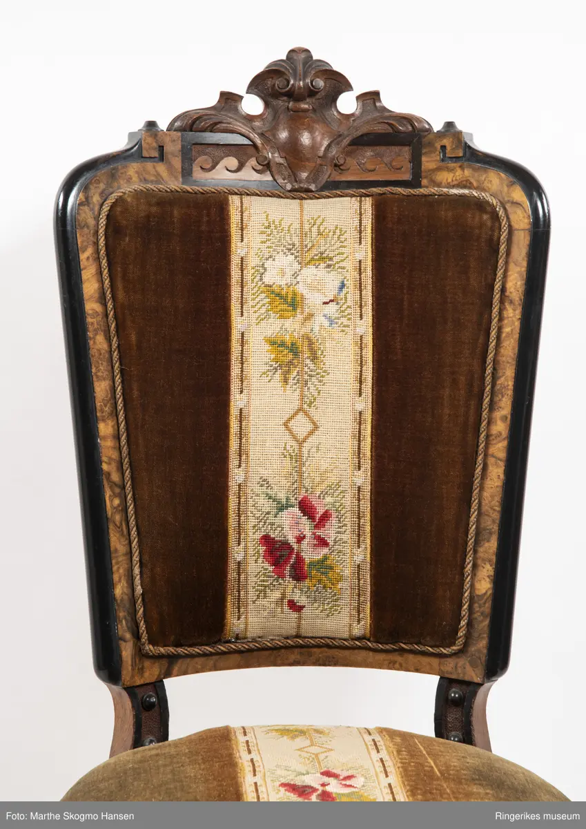Stol i brun mahogni med utskjæringer. Hjul på frembeina. Brunt fløyelstrekk på setet med korsstingsbroderi av blomster og geometriske mønster.