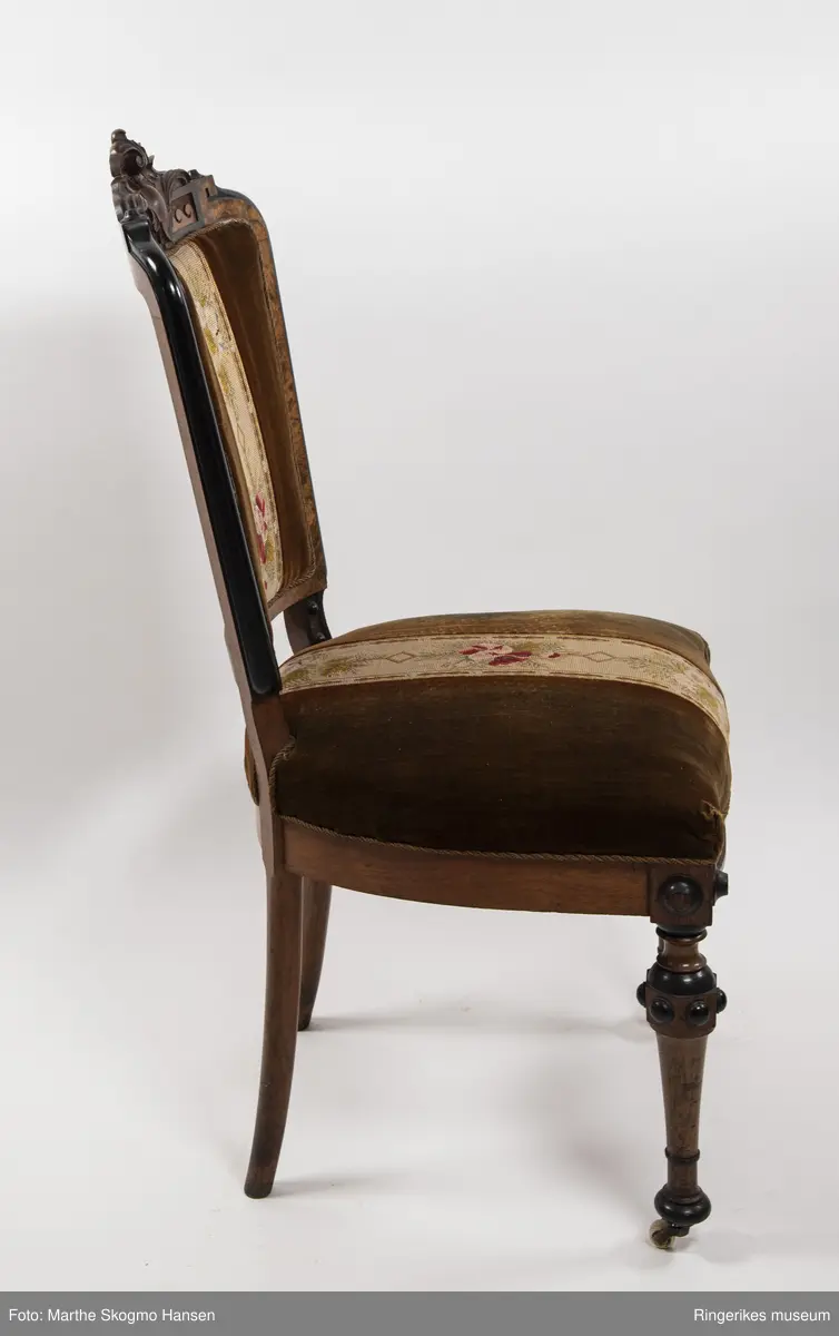 Stol i brun mahogni med utskjæringer. Hjul på frembeina. Brunt fløyelstrekk på setet med korsstingsbroderi av blomster og geometriske mønster.