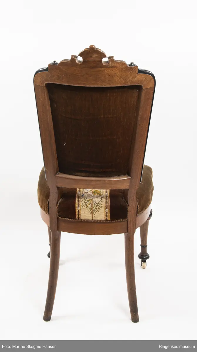 Stol i brun mahogni med utskjæringer. Hjul på frembeina. Brunt fløyelstrekk på setet med korsstingsbroderi av blomster og geometriske mønster.