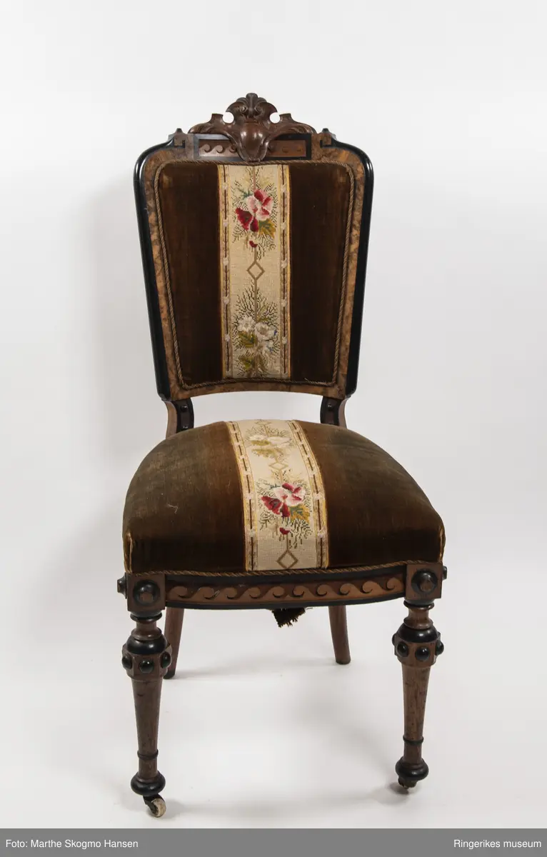 Stol i brun mahogni med utskjæringer. Hjul på frembeina. Brunt fløyelstrekk på setet med korsstingsbroderi av blomster og geometriske mønster.