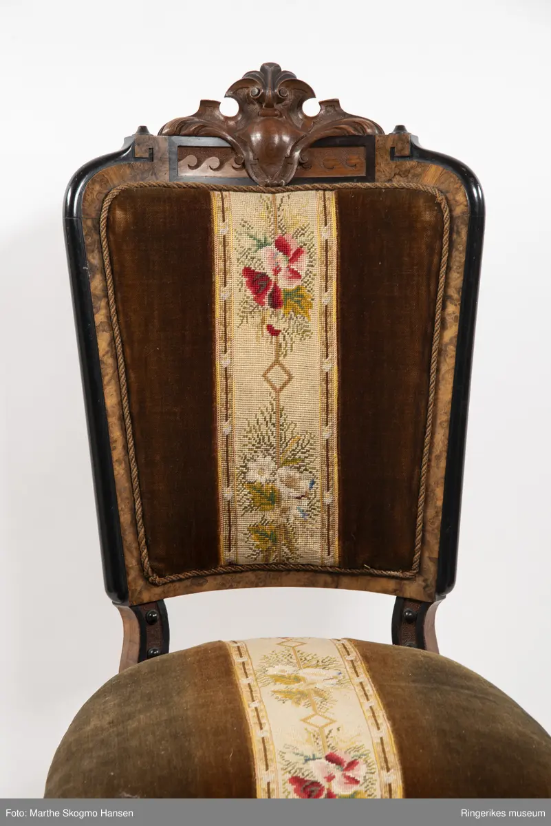 Stol i brun mahogni med utskjæringer. Hjul på frembeina. Brunt fløyelstrekk på setet med korsstingsbroderi av blomster og geometriske mønster.