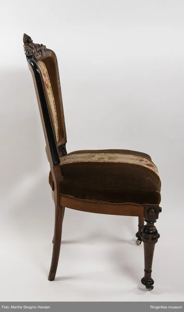 Stol i brun mahogni med utskjæringer. Hjul på frembeina. Brunt fløyelstrekk på setet med korsstingsbroderi av blomster og geometriske mønster.