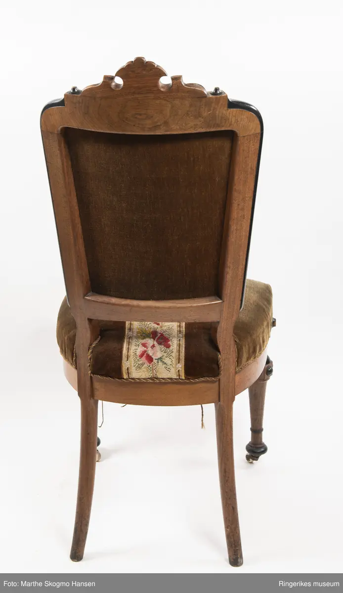Stol i brun mahogni med utskjæringer. Hjul på frembeina. Brunt fløyelstrekk på setet med korsstingsbroderi av blomster og geometriske mønster.