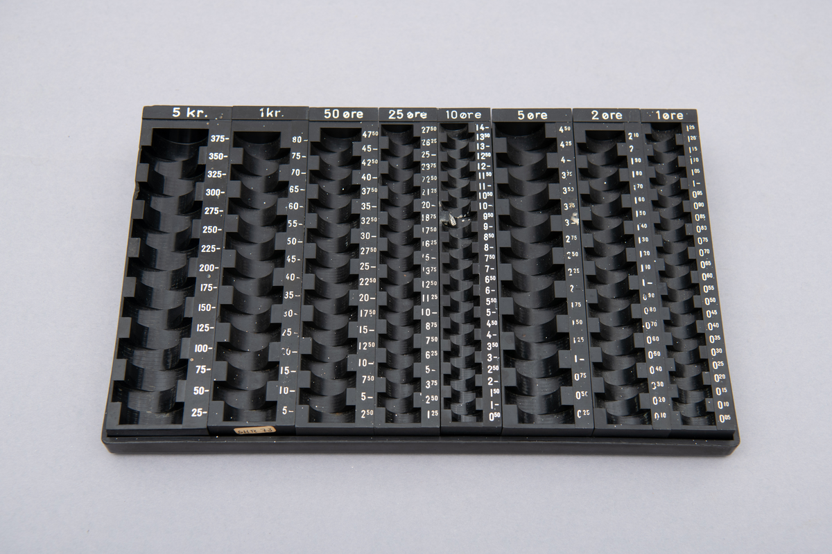 Myntkassett i svart bakelitt, inneholdende 8 behoholdere sortet på de ulike valørene 1, 2, 5, 10, 25 og 50 øre. 1 og 5 kroner. Hvit skrift.
