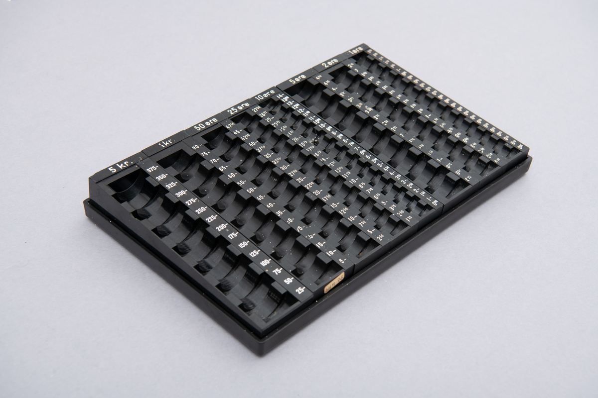 Myntkassett i svart bakelitt, inneholdende 8 behoholdere sortet på de ulike valørene 1, 2, 5, 10, 25 og 50 øre. 1 og 5 kroner. Hvit skrift.
