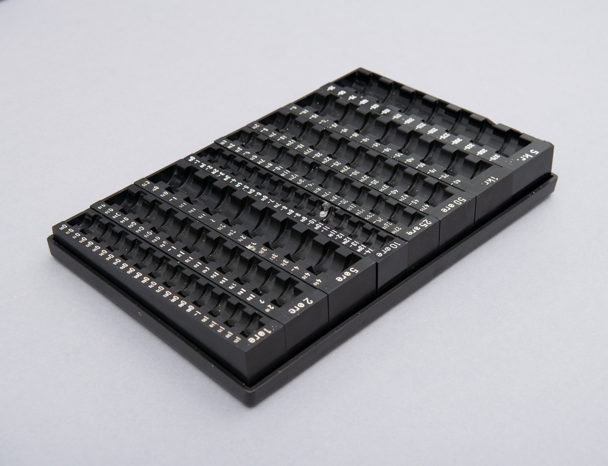 Myntkassett i svart bakelitt, inneholdende 8 behoholdere sortet på de ulike valørene 1, 2, 5, 10, 25 og 50 øre. 1 og 5 kroner. Hvit skrift.
