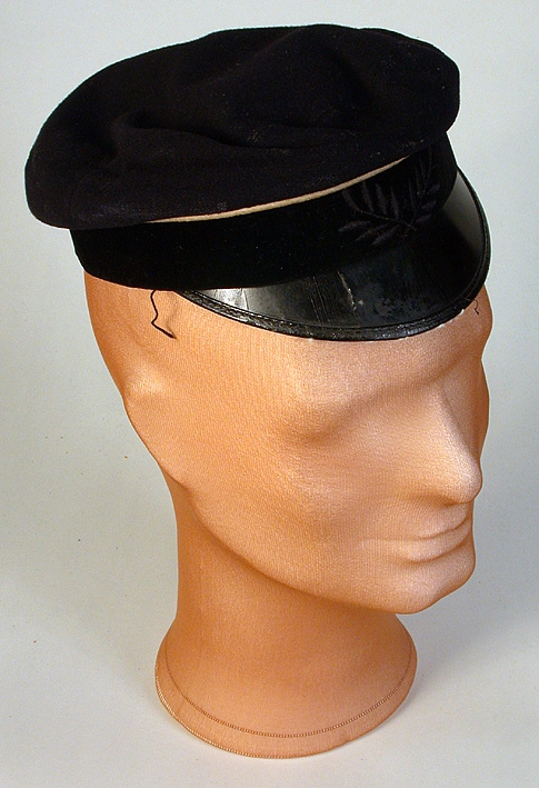 Sydd i svart ylle med lackerad skärm. Mösskanten med svart sammetsband med broderade blad mittfram.Vit snodd runt mössan. På kullen fastsydd en värmlandsörn av vitmetall. Svettsrem av brunt konstläder, fodrat med kyprat bomullstyg i gult och blått. På svettsremmen stansade små hål. P G R